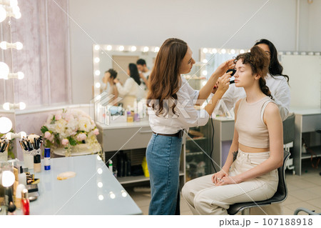 Wide shot of female hairdresser using hair tongs curling hair of young woman in beauty studio. Makeup artist applying foundation on lady face using sponge. Concept of make up and styling in 4 hands. 107188918