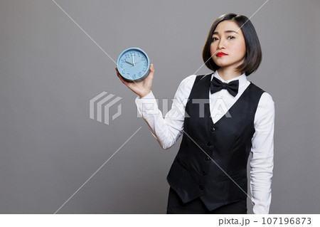 Hotel woman receptionist holding retro alarm clock and looking at camera with serious facial expression. Young asian waitress wearing uniform showing time management concept portrait 107196873