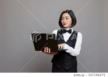 Young asian waitress using laptop, checking cafe online menu and managing catering service. Attractive restaurant receptionist wearing professional uniform and holding portable computer in studio 107196875
