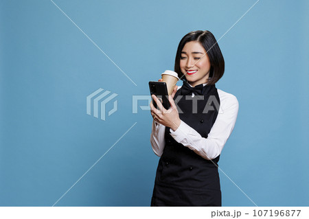 Smiling asian waitress wearing cafe uniform enjoying coffee to go and texting on smartphone. Cheerful woman receptionist holding tea paper mug and reading message on mobile phone 107196877