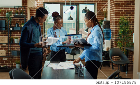 Team of people analyzing startup results in business meeting, discussing about executive data project. Colleagues examining analytics on papers and laptop, making decisions in office. 107196878