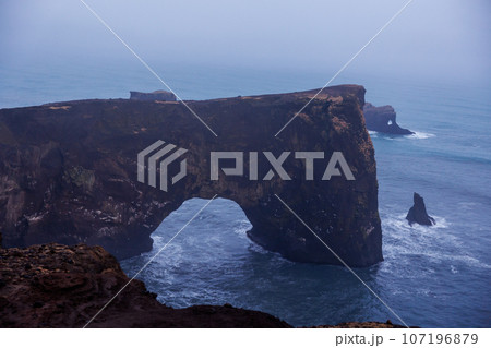 Rocky arch gate on nordic peninsula, dyrholaey natural landmark surrounded by beautiful scenery and stone hills. Massive coastline shore with rocks and fantastic structure, foggy bay. 107196879