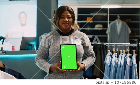 Young store manager showing tablet with green screen, advertising fashion boutique with chroma key display. Female employee using isolated mockup template with blank copyspace. 107196886