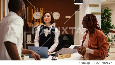 Friendly receptionist presenting holiday package options to african american couple in posh hotel. Husband and wife on honeymoon trip preparing to pay for extra room amenities in resort lounge Friendly receptionist presenting holiday package options to african american couple in posh hotel. Husband and wife on honeymoon trip preparing to pay for extra room amenities in resort lounge 107196937