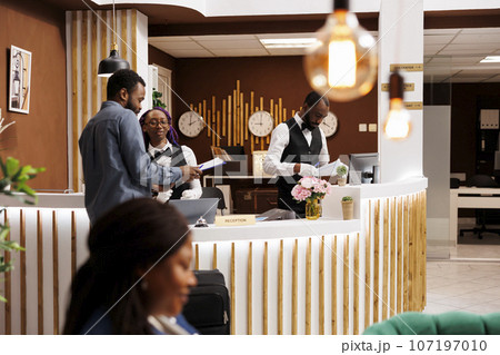African American man tourist standing at reception with luggage filling check-in form upon arrival at hotel. Black guy traveler signing documents at front desk while checking in African American man tourist standing at reception with luggage filling check-in form upon arrival at hotel. Black guy traveler signing documents at front desk while checking in 107197010