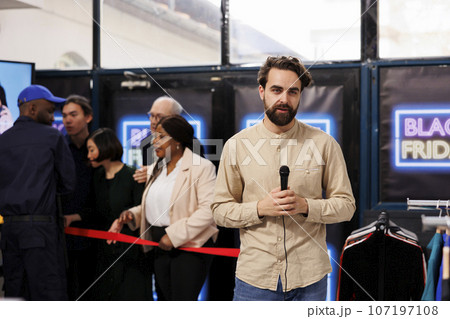Young man TV reporter holding microphone standing against eager crowd of shoppers at store entrance. Male journalist reporting on Black Friday shopping frenzy, making broadcast about seasonal sales Young man TV reporter holding microphone standing against eager crowd of shoppers at store entrance. Male journalist reporting on Black Friday shopping frenzy, making broadcast about seasonal sales 107197108