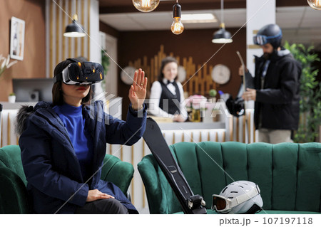 Asian lady wearing digital VR headset having skiing equipment and winter jacket in hotel lobby. Female tourist searching with vr glasses for best slopes at ski mountain resort snow adventure. 107197118