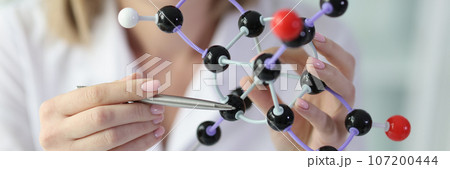 Female scientist holds model of molecular structure and points to one molecule close-up. 107200444