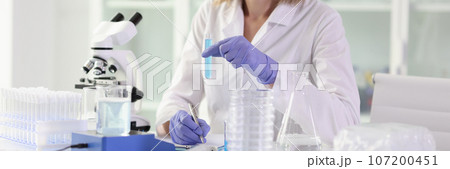 Smiling female scientist holding test tube with blue liquid and making notes. 107200451