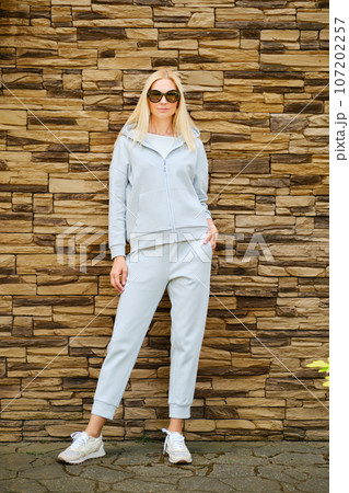 Blonde woman in tracksuit and sneakers by the wall 107202257