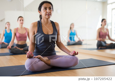 Concentrated Latina sitting in lotus pose with group of women Concentrated Latina sitting in lotus pose with group of women 107205422