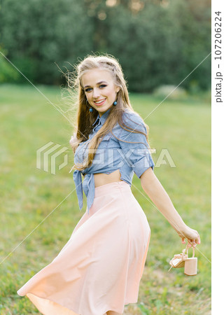 Young Caucasian woman or girl in a summer dress and a denim shirt is walking on the grass, holding shoes in her hands, relaxing outside the city on a weekend in summer Young Caucasian woman or girl in a summer dress and a denim shirt is walking on the grass, holding shoes in her hands, relaxing outside the city on a weekend in summer 107206224