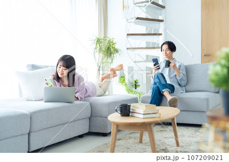 パソコンとスマホを見るリビングでくつろぐ若いカップル パソコンとスマホを見るリビングでくつろぐ若いカップル 107209021