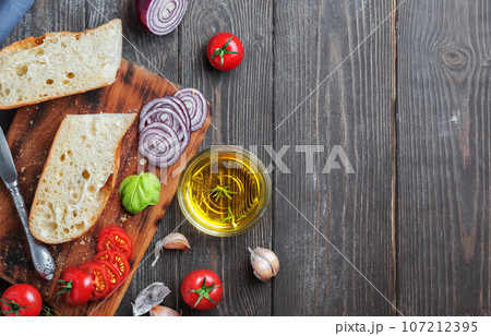 Fresh baguette sliced into pieces, tomatoes, vegetables and olive oil, ingredients for making a vegan sandwich, close-up. Dark wooden background, copy space for text 107212395