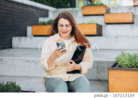 Elegant young 30s woman formally dressed, sitting on the bench and holding a folder with documents outdoors. Businesswoman with smart phone standing at the city. Elegant young 30s woman formally dressed, sitting on the bench and holding a folder with documents outdoors. Businesswoman with smart phone standing at the city. 107213189