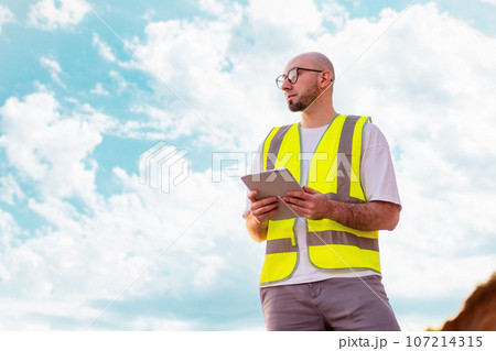 Clear air. Bottom view of bald bearded man with glasses holding tablet against blue cloudy sky. Copy space. Concept of environmental protection and recycling industry 107214315