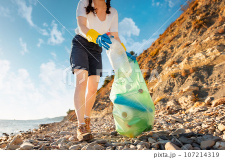 Bottom view of Young woman in rubber gloves collects plastic bottle on pebble wild beach. Concept of ecological disaster 107214319