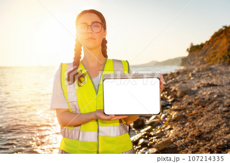 Inspection environmentalist and analyzing water in ocean. Volunteer show tablet's screen with mock up. In background is ocean, coast and sunset. Concept of environment pollution 107214335
