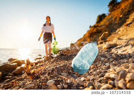 Defocused view of woman volunteer holds full plastic bag of trash and walking down of pebble wild beach. Close up of plastic bottle in foreground. Concept of protection ocean and Earth Day 107214338
