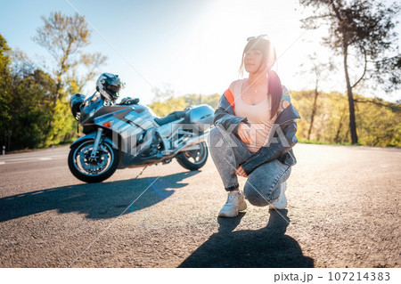 World Motorcyclist Day. A young woman with tattoos on her arm, wearing leather jacket, and posing sitting. Road and motorbike in the background. Motorcycle local travel concept World Motorcyclist Day. A young woman with tattoos on her arm, wearing leather jacket, and posing sitting. Road and motorbike in the background. Motorcycle local travel concept 107214383