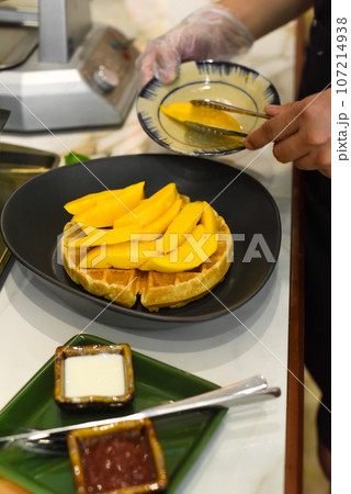 the cook putting fresh sliced mango fruit on baked belgian waffles with tongs 107214938