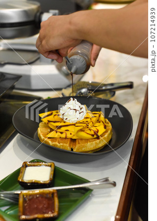 the cook putting chocolate syrup on fresh sliced mango fruit on baked belgian waffles the cook putting chocolate syrup on fresh sliced mango fruit on baked belgian waffles 107214939