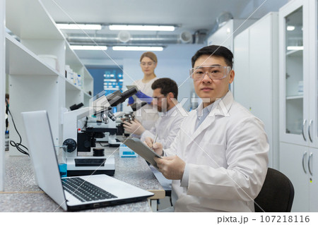 International group of young scientists work in the laboratory. An Asian man is sitting at a desk and using a tablet, behind which colleagues are examining with a microscope. 107218116