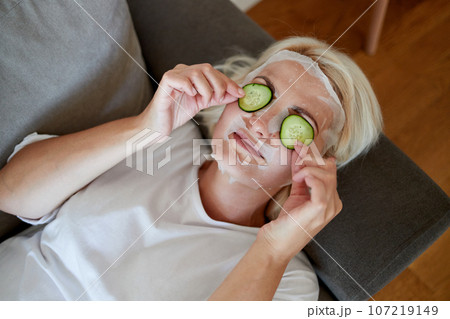 Blonde woman with a fabric mask on her face and cucumber slices on her eyes 107219149