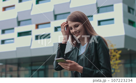 Young Attractive Girl with Long Brunette Hair walking in the Street, Having Mobile Phone in her Hands. Typing Messages and Smiling while sending. Having fashionable clothes 107223234