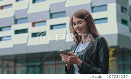 Young Attractive Girl with Long Brunette Hair walking in the Street, Having Mobile Phone in her Hands. Typing Messages and Smiling while sending. Having fashionable clothes Young Attractive Girl with Long Brunette Hair walking in the Street, Having Mobile Phone in her Hands. Typing Messages and Smiling while sending. Having fashionable clothes 107223237