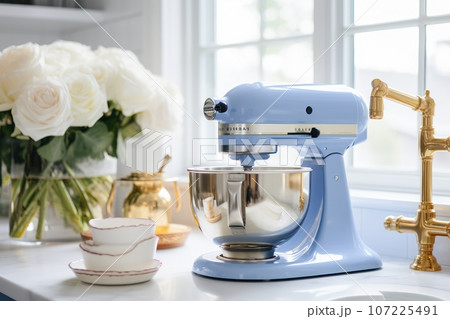 Fragment of white kitchen with a window and golden faucet on the background. Blue planetary mixer with stainless steel bowl, cups with ingredients and bouquet of white roses in a glass vase. Close-up. 107225491