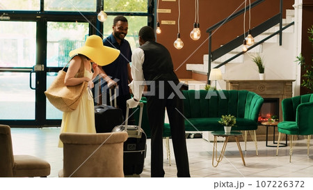 Hotel bellhop providing luxury service to guests entering reception lobby, concierge carrying baggage and trolley bags to room. Young couple arriving at tropical resort front desk. 107226372