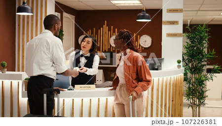 Friendly bellhop employee showing tourists the way to their room while carrying luggage after check in process is over. Stressed hotel manager verifying guests booking paperwork 107226381