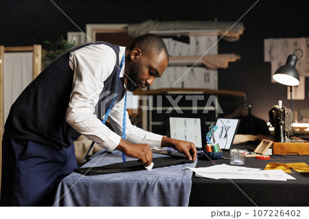 Tailor creating sophisticated suit blazer surrounded by sewing instruments and luxurious fabrics in atelier shop workspace. Couturer finishing business attire for upcoming sartorial comission 107226402