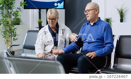 Senior medic measuring patient hypertension using tonometer instrument, checking hypotension during healthcare appointment in hospital waiting room. Elderly man having cardiac exam Senior medic measuring patient hypertension using tonometer instrument, checking hypotension during healthcare appointment in hospital waiting room. Elderly man having cardiac exam 107226407