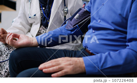 Elderly medic using medical tonometer to measure patient pulse pressure and hypertension, doing cardiology examination in hospital waiting room. Senior man attending checkup visit. Medicine concept Elderly medic using medical tonometer to measure patient pulse pressure and hypertension, doing cardiology examination in hospital waiting room. Senior man attending checkup visit. Medicine concept 107226408