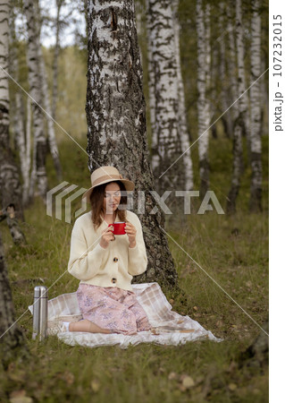 A young beautiful woman in a dress and a round hat reads a book outdoors in the forest and drinks tea. Romantic and vintage photo of a beautiful girl. Reading and relaxation A young beautiful woman in a dress and a round hat reads a book outdoors in the forest and drinks tea. Romantic and vintage photo of a beautiful girl. Reading and relaxation 107232015