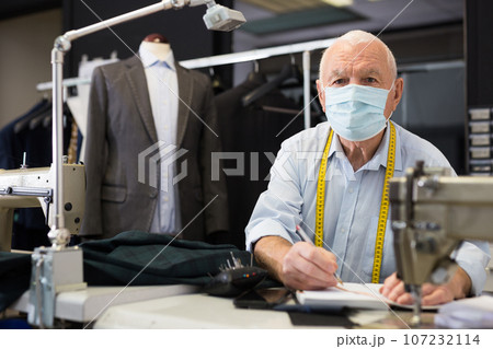 Portrait of an elderly tailor in protective mask making notes in notebook 107232114