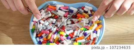 Women's hands hold knife and fork over heart-shaped plate with bunch of different pills. Women's hands hold knife and fork over heart-shaped plate with bunch of different pills. 107234635