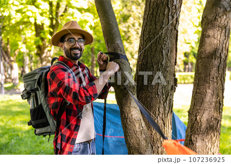 Smiling dark-haired bearded young traveler standing near the tree Smiling dark-haired bearded young traveler standing near the tree 107236925