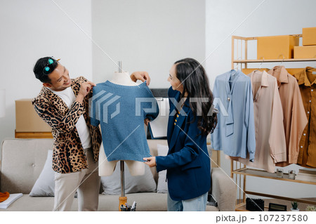 Couple of fashion designers working with fabric and clothing sketches at the studio full of tailoring tools and holds tablet and laptop. 107237580