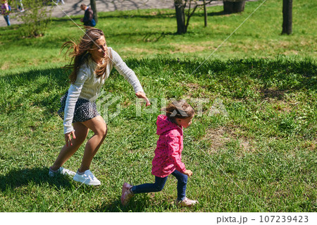 A young mother catches her child, little daughter playing on the green grass A young mother catches her child, little daughter playing on the green grass 107239423