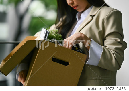 Upset female office worker carrying cardboard box with belongings, leaving office after quitting job. Unemployed concept Upset female office worker carrying cardboard box with belongings, leaving office after quitting job. Unemployed concept 107241878