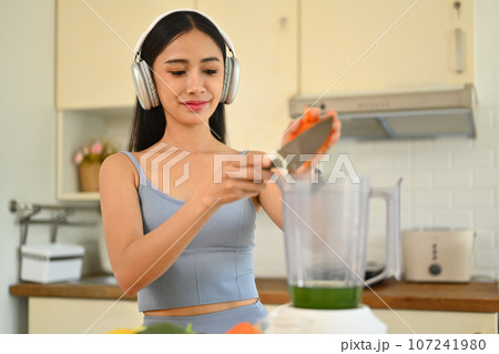 Young fitness woman in headphone trying new recipes for green detox vegetable smoothie. Healthy eating concept 107241980