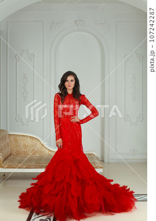 Beautiful brunette woman in red dress standing in luxurious white interior Beautiful brunette woman in red dress standing in luxurious white interior 107242287