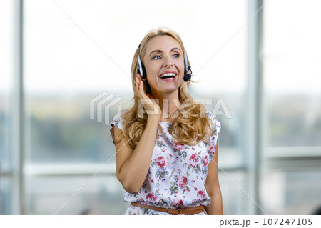 Portrait of smiling blond woman wearing headset indoors. Blurred window in the background. Portrait of smiling blond woman wearing headset indoors. Blurred window in the background. 107247105