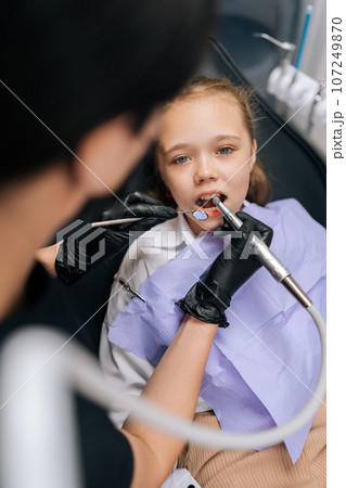 Vertical portrait of adorable little child girl sitting in stomatology seat with open mouth looking at camera while unrecognizable orthodontist examining teeth, with dental tool in dentistry clinic. Vertical portrait of adorable little child girl sitting in stomatology seat with open mouth looking at camera while unrecognizable orthodontist examining teeth, with dental tool in dentistry clinic. 107249870