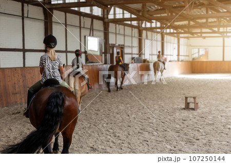 Horse riding school. Little children girls at group training equestrian lessons in indoor ranch horse riding hall. Cute little beginner blond girl kid in helmet sitting on brown horse horseback 107250144
