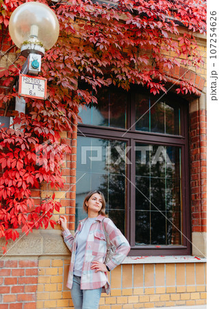 a beautiful girl stands against the background of the window of an old European house, entwined with a floating red color in autumn. Tourism & Travel Concept. Nice portrait of a young woman, in boho 107254626