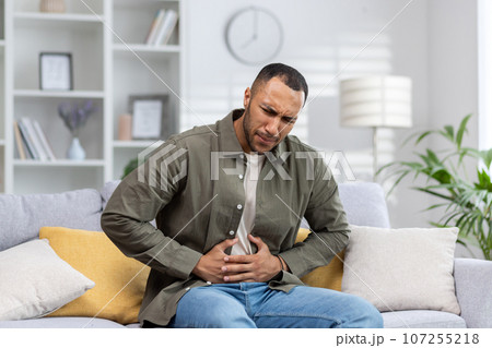 A young Muslim man grimaced in pain and holds his hands to the side of his stomach, suffering from appendicitis, convulsions. Sitting at home on the sofa, waiting for emergency medical help. 107255218